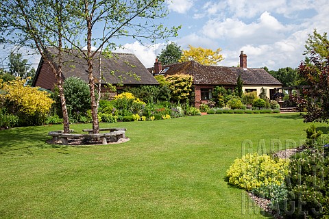 Sweeping_lawns_and_borders_of_herbaceous_perennials_shrubs_and_mature_tree_with_wooden_seat_around_i